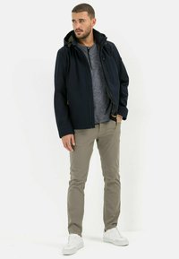 Veste à capuche bleu marine en tissu doux, avec fermeture éclair sur le devant et matériel doré, associée à une chemise henley grise et un pantalon beige clair. Baskets blanches.