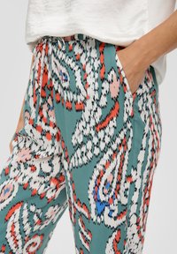 Wielokolorowe spodnie w wzory na turkusowym tle, z abstrakcyjnymi wzorami w kolorach czerwonym, różowym i niebieskim. Elastyczna talia ze sznurkiem i boczne kieszenie.