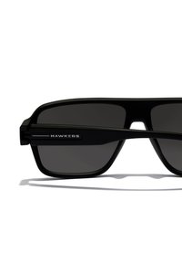 Mustad ristkülikukujulised päikeseprillid tumedate läätsedega, mattviimistlusega ja stiilse disainiga. Bränd "HAWKERS" on nähtav prillide küljel.