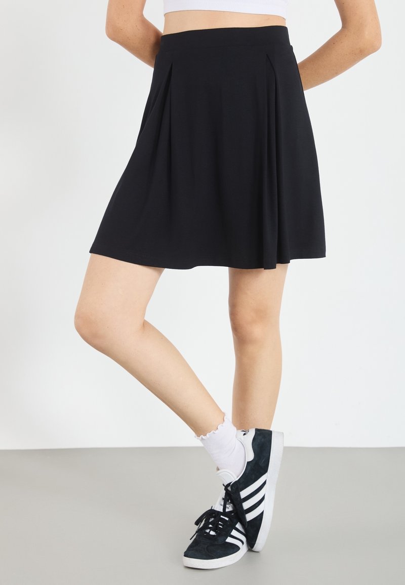 Zwarte geplooide rok met een lengte tot halverwege de dij, gemaakt van zachte stof; gecombineerd met zwarte sneakers met witte strepen en enkellange witte sokken.