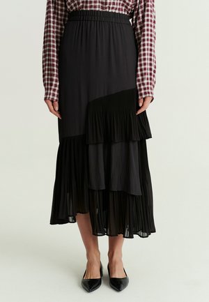 Femme portant une jupe midi plissée noire avec des détails superposés, associée à une chemise à carreaux rouge et blanche à manches longues et des chaussures noires pointues.