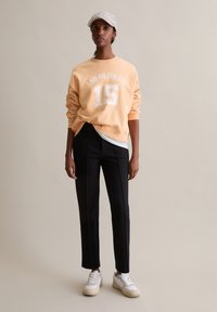 Brzoskwiniowy sweatshirt z napisem "JESTEM SPORTOWCEM 15" w kolorze białym, w połączeniu z czarnymi eleganckimi spodniami i białymi sneakersami. Dodatkowo szara czapka w wzory.