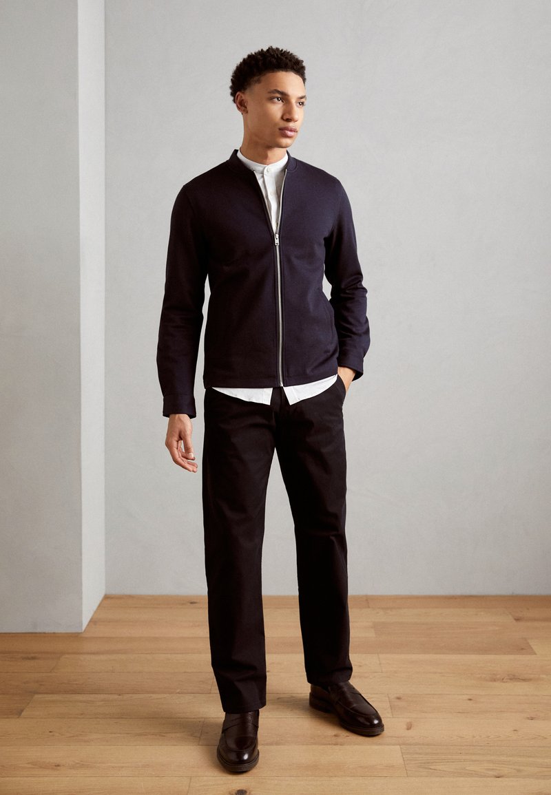 Veste zippée marine à texture lisse, associée à une chemise blanche à col, un pantalon noir et des chaussures en cuir marron foncé, debout sur un sol en bois.