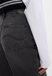 Gros plan d'une personne portant un jean Lee gris foncé avec une poche arrière visible et une manche de chemise blanche à manches longues couvrant une partie de la main.