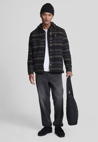 Camicia di flanella a quadri neri e oro, T-shirt bianca, jeans grigi, sneakers nere e borsa a tracolla nera, modello in posa contro uno sfondo semplice.