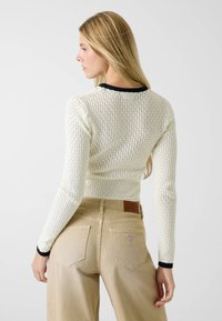 Gebreide trui in crème met open lattice-patroon en zwarte geribbelde halslijn. Gecombineerd met beige broek met hoge taille en achterzakken.