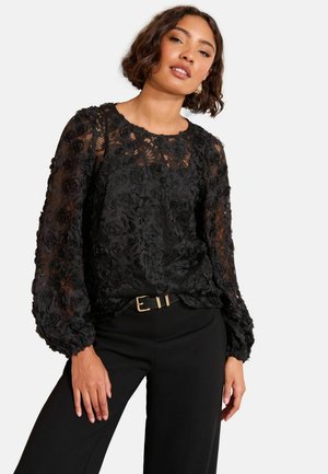 Schwarze Bluse aus floraler Spitze mit langen Puffärmeln, rundem Ausschnitt und strukturierten Rosenmustern, kombiniert mit hochgeschnittenen schwarzen Hosen.