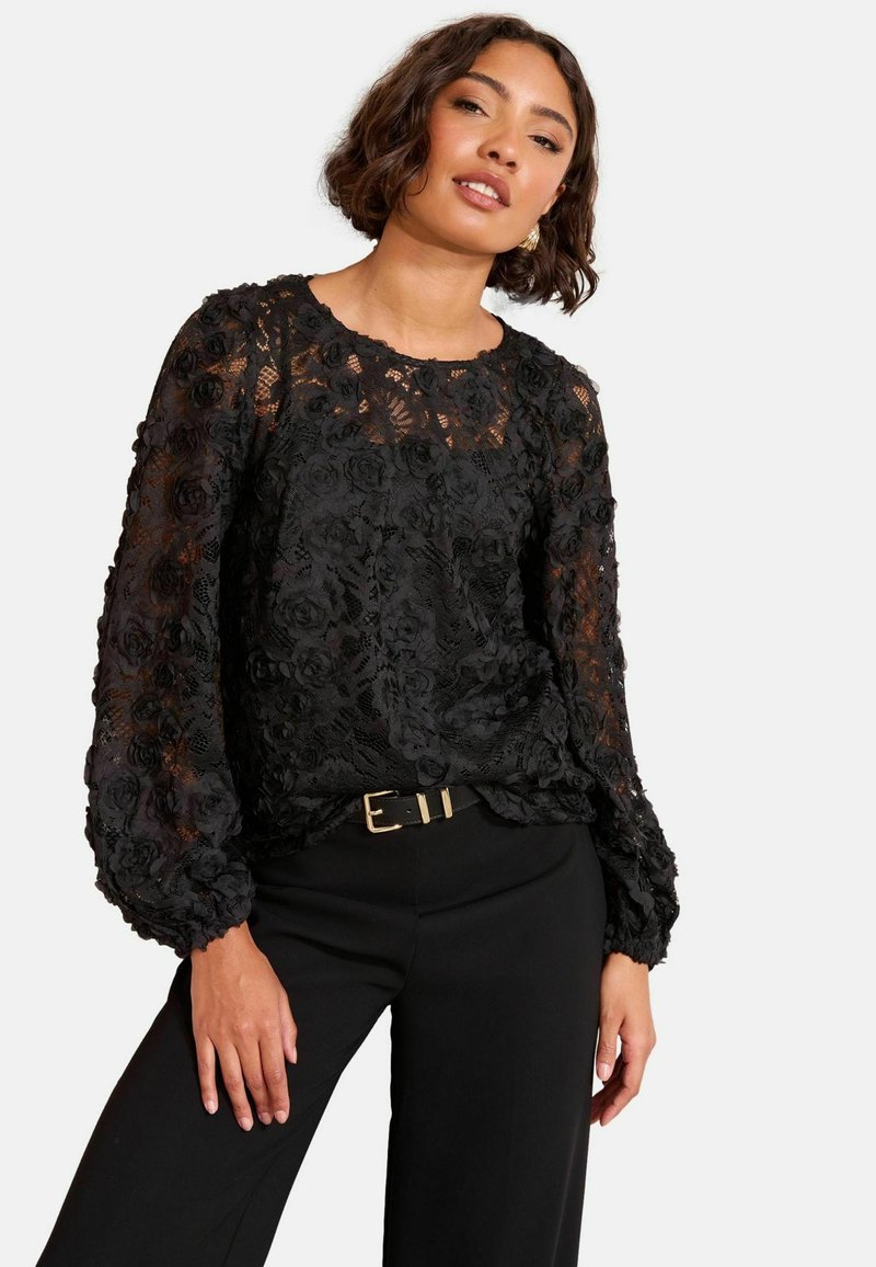 Črna cvetna čipkasta bluzica z dolgimi napihnjenimi rokavi, okroglo izrezom in teksturiranimi rožnatimi vzorci, v kombinaciji z visokim pasom in črnimi hlačami.