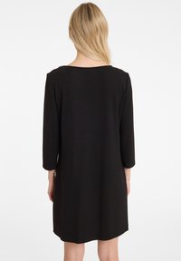 Robe noire mi-longue avec des manches trois-quarts, un col rond et un tissu lisse. Le dos présente un design simple et minimaliste.