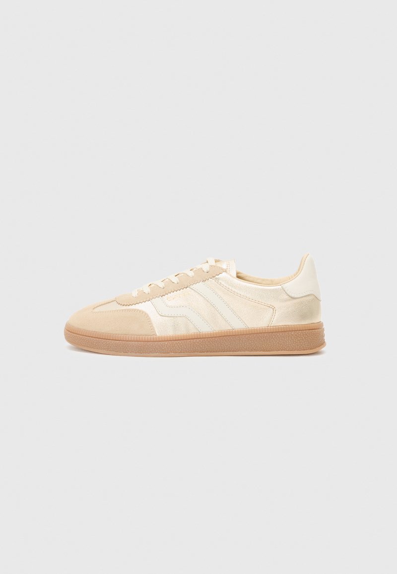 Sko med en beige overdel som har semsket og glatte teksturer. Tre hvite striper langs siden og en gummisåle.