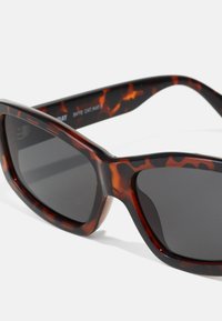 Gafas de sol carey con lentes oscuros, un diseño envolvente y ancho, y montura de plástico suave con un acabado brillante y acentos contrastantes.