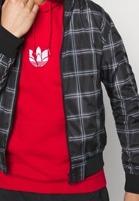 Zwarte geruite bomberjack met witte lijnen over een rode hoodie. Kenmerken zijn ribbelmanchetten en -zoom, ritssluiting en zakdetails.