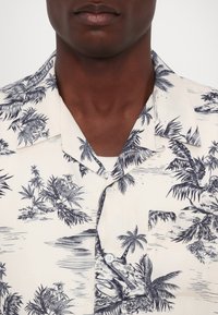 Short-sleeve button-up shirt featuring a light background with dark blue palm tree and floral patterns, a chest pocket, and a classic collar.