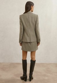 Woman standing facing away wearing a houndstooth blazer and matching skirt with black knee-high heeled boots on a neutral background.