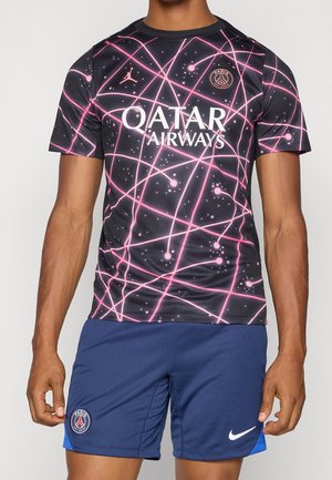 Homme portant le maillot de football du Paris Saint-Germain avec un motif abstrait noir et rose et un short bleu avec les logos du club et de Nike.