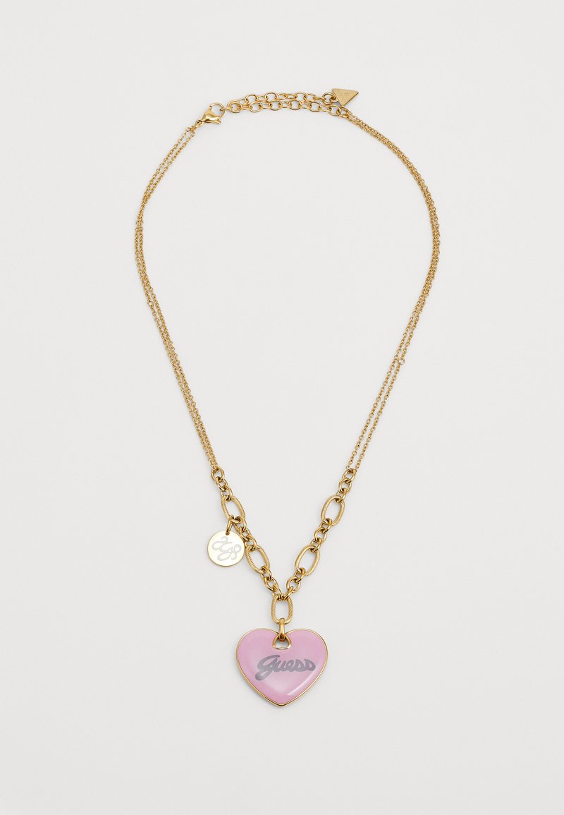 Gullfarget kjedehalskjede med lenkedesign, en rosa hjertecharme med "Guess" i grått, og en liten rund etikett.