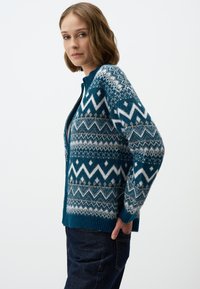 Cardigan en tricot teal avec un motif géométrique en blanc et beige. Il présente une fermeture éclair et des poignets côtelés, offrant une apparence texturée.