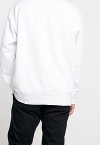 Felpa bianca con un fit rilassato, caratterizzata da maniche lunghe, orlo arrotondato e polsini a coste. Indossata sopra pantaloni neri, che mettono in mostra un design minimalista.