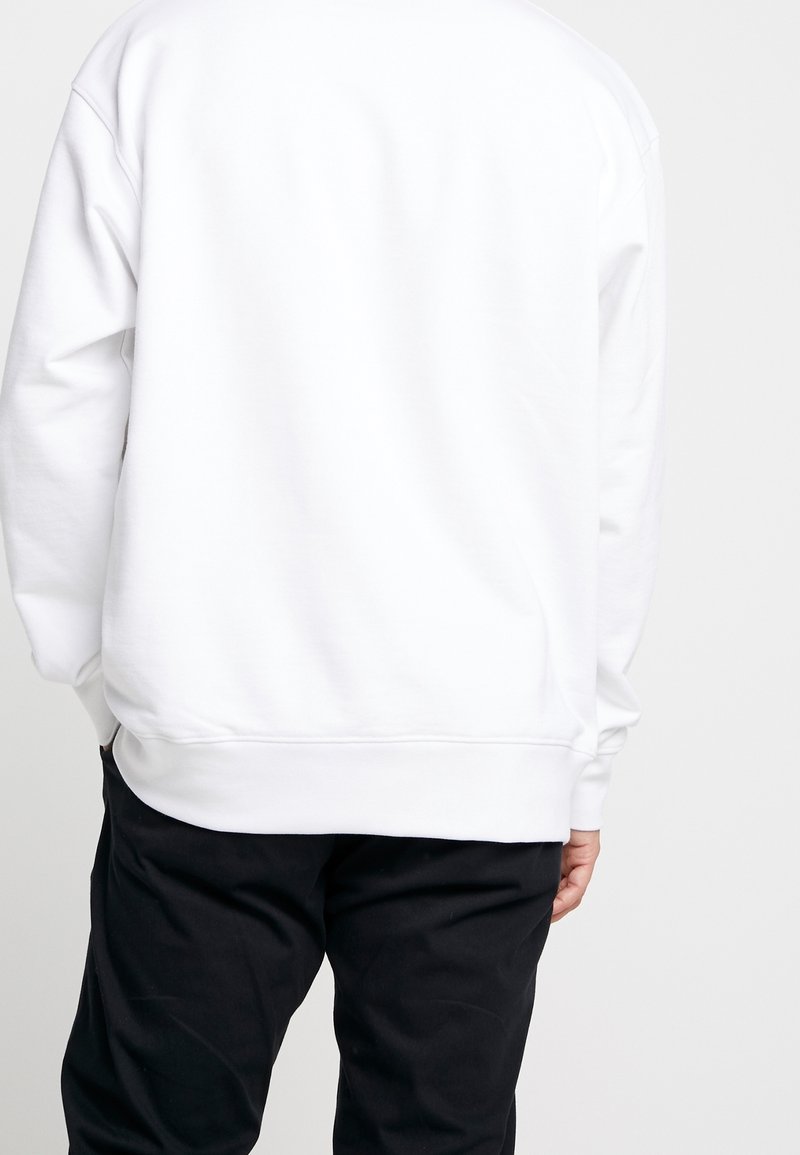 Felpa bianca con un fit rilassato, caratterizzata da maniche lunghe, orlo arrotondato e polsini a coste. Indossata sopra pantaloni neri, che mettono in mostra un design minimalista.