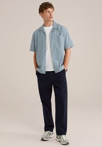 Camicia a maniche corte a righe blu chiaro, t-shirt bianca a girocollo sotto, abbinata a pantaloni blu scuro e sneaker grigie con dettagli verdi.