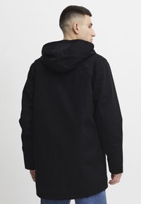 Sort hættejakke i en afslappet skæring med en glat tekstur og forlommer, båret over lyse denim. Synlig syning.