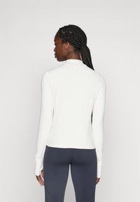 Vrouw met gevlochten haar die een lange mouw witte top en donkere grijze leggings draagt, met haar rug naar de camera gekeerd tegen een effen lichte achtergrond.