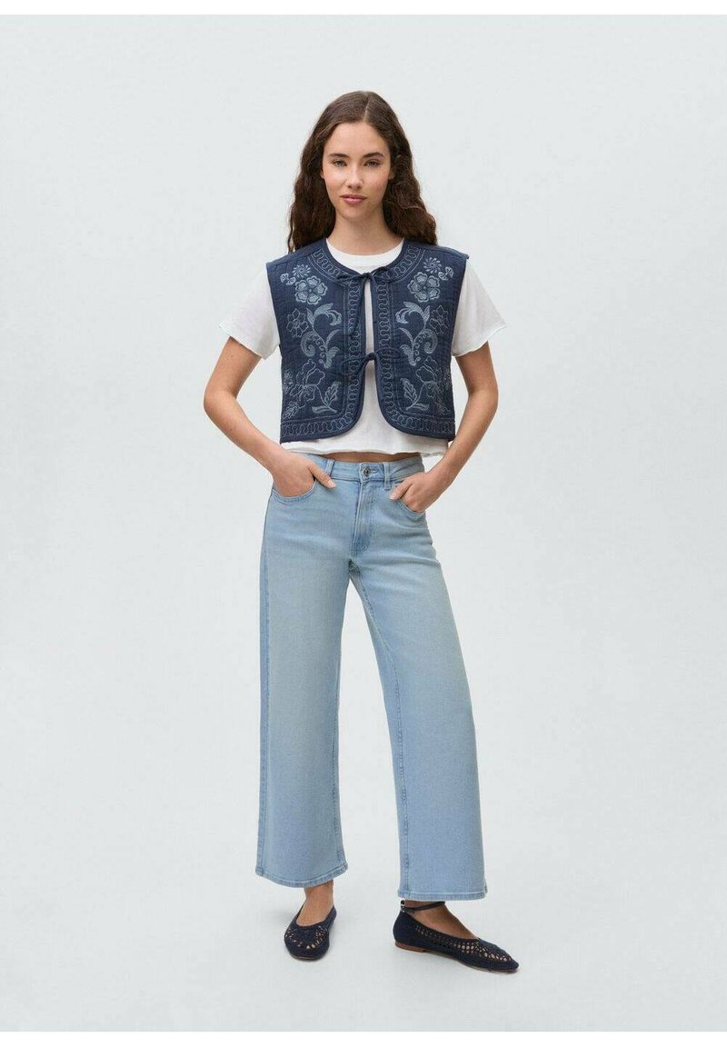 Mornarsko modro floralno vezeno telovnik s okroglim izrezom, oblečen preko bele majice, kombiniran s svetlo modrimi širokimi hlačami in temnimi balerinkami.