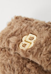 Rjava torbica iz umetnega krzna z mehkobno teksturo in izrazitim zlatim logotipom na sprednji strani. Kompaktna in pravokotna oblika.