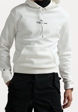 Mężczyzna w białej bluzie z kapturem Polo Ralph Lauren z przednią kieszenią i czarnych spodniach typu cargo, ręce częściowo w kieszeniach, na jednolitym tle.