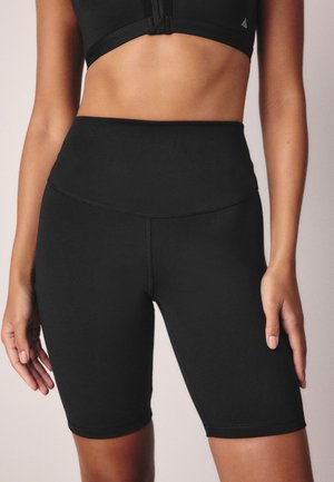 Kvinde iført sorte cykelshorts med høj talje og en sort kort sportstop, stående med armene afslappet ned langs siden.