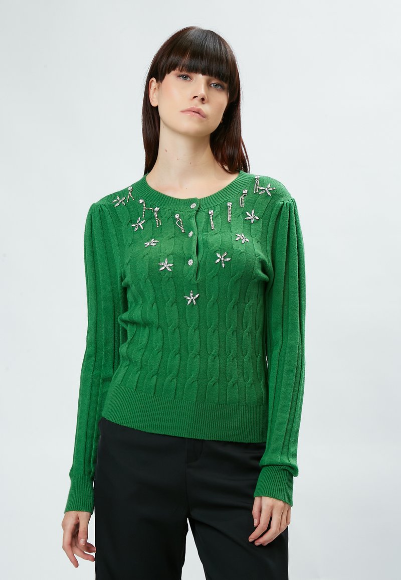 Grüner Kabelstrickpullover mit langen Ärmeln, verziert mit floralen Akzenten und einem runden Ausschnitt. Das Material ist strukturiert und hat gerippte Bündchen.