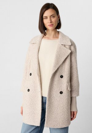 Lichtbeige dubbelzijdige jas van een textuurstof met grote zwarte knopen, gelapte kraag en zijzakken. Gedragen over een crèmekleurige trui.
