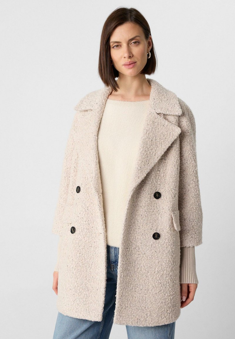 Hellbeige doppelt knöpfte Jacke aus strukturierter Stoff mit großen schwarzen Knöpfen, Reverskragen und seitlichen Taschen. Getragen über einem cremefarbenen Pullover.