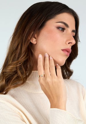 Anello d'oro con due pietre quadrate, indossato su una mano. Sullo sfondo si vede un'illuminazione chiara, mentre il modello ha lunghi capelli mossi. Tessuto strutturato di colore crema.