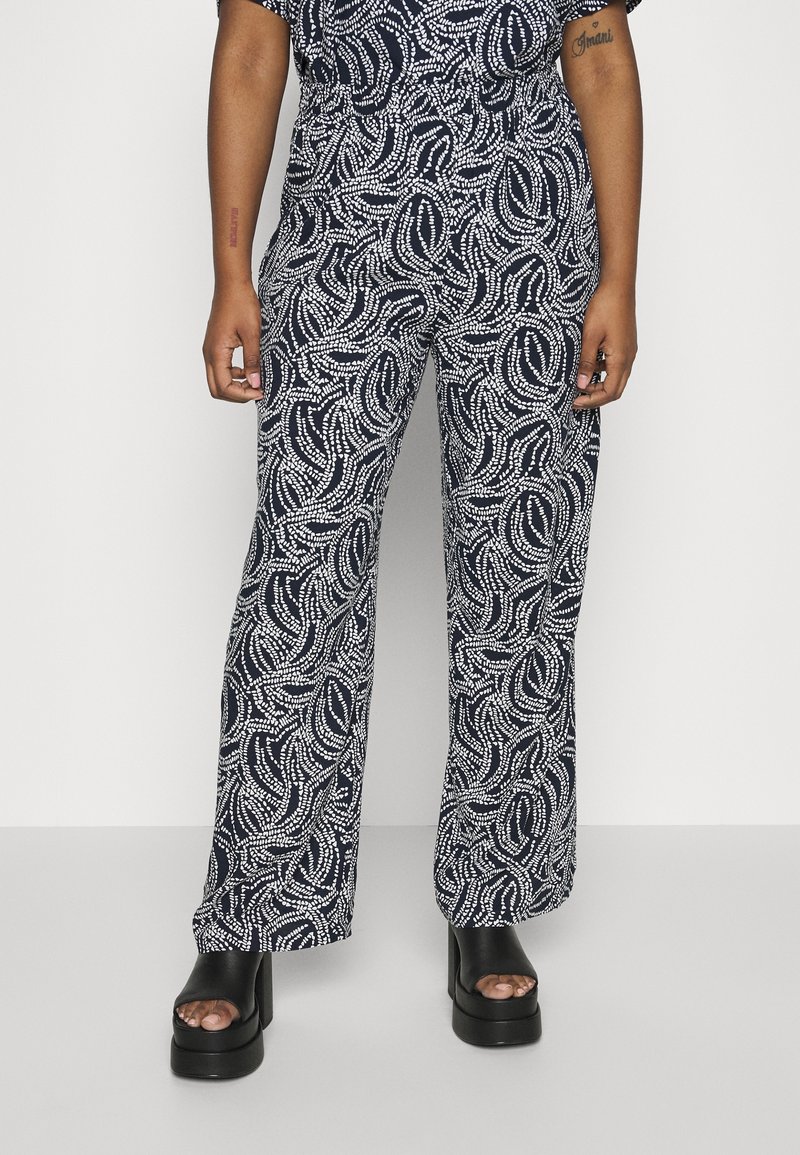 Personne portant un pantalon à motif abstrait noir et blanc et des chaussures plates-formes noires à bout ouvert, debout devant un fond blanc uni.