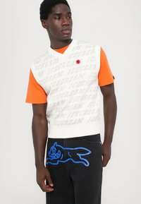 Jersey sin mangas blanco con texto gris "ICECREAM" y logo rojo. Camiseta naranja debajo. Pantalones vaqueros negros con un gráfico de dibujos animados azul en la pierna.