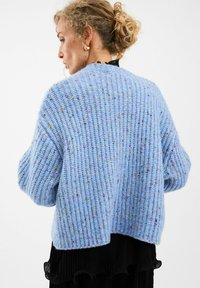 Svetlo modrý pletený kardigán s rebrovanou textúrou, ktorý má farebné škvrny po celom povrchu. Voľný strih s spadnutými ramenami a skráteným zadným dielom.