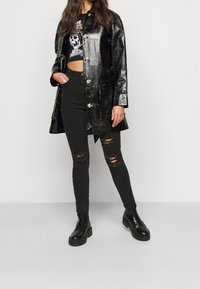 Abrigo de cuero sintético negro y brillante con textura de cocodrilo, con grandes botones y bolsillos laterales, llevado sobre una camiseta gráfica negra de corte cropped y jeans rasgados negros.