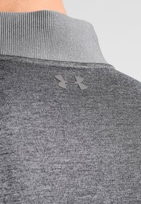 Primer plano de un tejido gris texturizado en la parte superior de la espalda y el cuello de una prenda que presenta el logo de Under Armour.