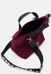 Vínově červená látková taška s černými koženými rukojeťmi a nastavitelným popruhem. Obsahuje zapínání na zip a logo na straně. Vnitřek je černý.