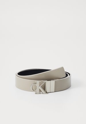 Ceinture en cuir beige avec une texture lisse et une boucle en métal argenté poli ornée du logo "CK". Le design est minimaliste et épuré.