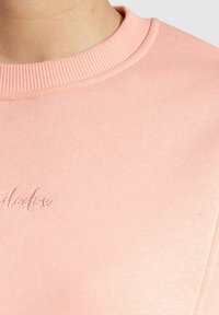 Lichtroze sweatshirt met een geribde ronde hals en geborduurd logo op de voorkant. De stof lijkt zacht met een gladde textuur.