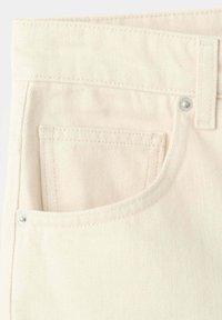 Gros plan d'une poche de jean en denim couleur crème avec des coutures visibles et des rivets argentés sur la ceinture et le coin de la poche.