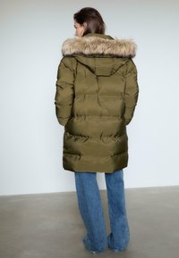 Oliwkowa kurtka puchowa z kapturem wykończonym futrem, o pikowanym wzorze i średniej długości sylwetce, zestawiona z szerokimi dżinsami.