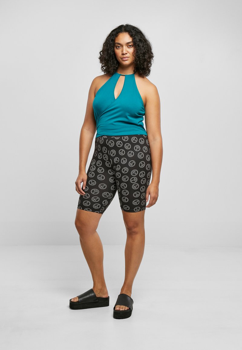 Teal ermeløs topp med nøkkelhull-design, paret med svarte sykkelshorts med et hvitt bladmønster og kraftige svarte sandaler.
