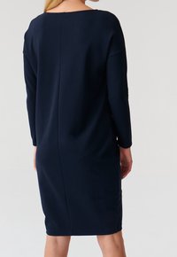 Mörkblå långärmad klänning med vid halsringning, figursydd silhuett och längd vid vadarna. Har ett mjukt tyg med en subtil lyster.