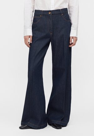 Person iført mørkeblå vidde jeans med forlommer og en hvid skjorte, sorte sko synlige under jeansene.
