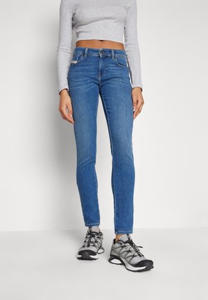 Jeans skinny de mezclilla azul con cinco bolsillos, trabillas para cinturón y detalles de costura sutiles, combinados con zapatillas deportivas grises y una camiseta gris claro.