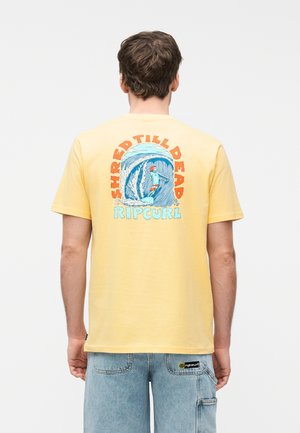 Man draagt een geel T-shirt met een kleurrijke surfprint en de tekst "Shred Till Dead Rip Curl" op de achterkant, gecombineerd met lichtblauwe spijkerbroek.