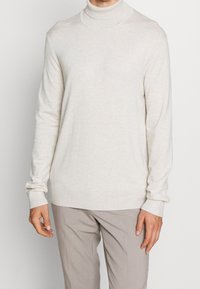 Ett krämfärgat polokrage-sweater gjort av mjukt stickat material. Har en figurnära design, ribbade manschetter och en slät textur. Bärs tillsammans med beige byxor.